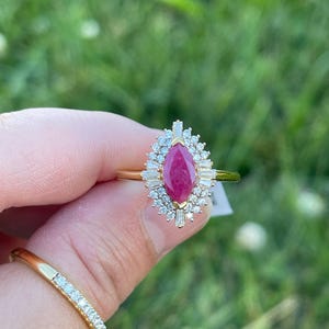 May include: A gold ring featuring a central marquise-cut pink gemstone surrounded by a halo of small round and baguette-cut white stones. A second thin gold band with a row of small white stones is visible below the main ring setting.