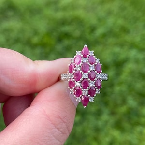 May include: A silver-toned ring featuring a cluster of oval-cut deep pink rubies arranged in a marquise-shaped setting, accented with small clear round stones between the rubies and along the band, creating a multi-stone floral-like design.