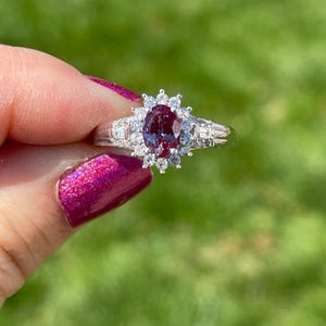 May include: A silver-toned ring featuring a central oval-cut deep red gemstone surrounded by a halo of small round clear stones. The band includes additional small clear stones set along the shoulders, creating a cluster design on a polished metal band.