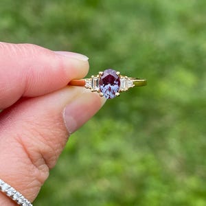 May include: A gold ring featuring a central oval-cut alexandrite gemstone that displays a purple and teal color shift. The center stone is flanked by two baguette-cut clear stones and two smaller round clear stones set in a gold band.