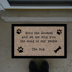 May include: A brown doormat with black text that reads "Ring the doorbell and let me sing you the song of my people - The Dog". The doormat has black paw prints and a bone design.