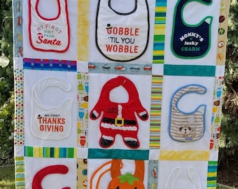 May include: A handmade baby bib quilt with various bibs featuring holiday and special occasion themes. Bibs include phrases like "My First Visit From Santa," "Gobble 'Til You Wobble," and "Mommy's Lucky Charm." The quilt is displayed outdoors.