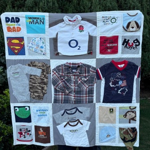 May include: A handmade quilt featuring squares of various children's clothing items. The quilt includes shirts and onesies with text and graphics, such as "DAD thinks I'm RAD", a Superman logo, and a shirt with the text "US". The quilt is a mix of colors.