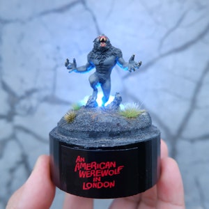 May include: A gray werewolf figurine with red eyes and a blue light base. The base is black and has the text "An American Werewolf in London" printed on it.