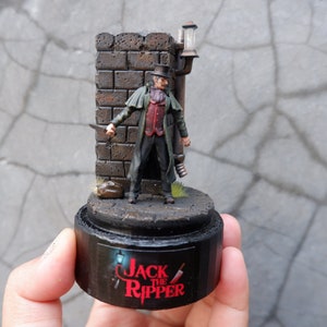 May include: A painted miniature figure of Jack the Ripper, standing in a dark alleyway with a brick wall and a street lamp. The figure is wearing a dark coat and hat, and holding a knife. The base of the miniature is painted black and has the words "Jack the Ripper" written on it.