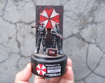 May include: A black and red Umbrella Corporation logo with a red biohazard symbol. Two black figures with guns stand in front of the logo. The figures are holding a black case with a red biohazard symbol. The logo is on a black base with the text "PROPERTY OF UMBRELLA CORPORATION" and a barcode.