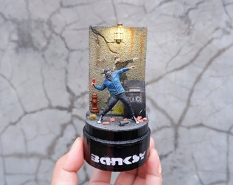May include: A small, detailed figurine of a masked figure throwing a fire extinguisher at a wall with the word "Police" on it. The figurine is on a black base with the word "Banksy" printed on it.