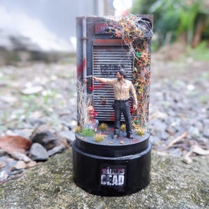 May include: A miniature diorama featuring a scene from "The Walking Dead." It depicts a figure in a tan shirt and dark pants, pointing a gun. The scene includes a building facade with a metal door, and the base reads "WALKING DEAD."