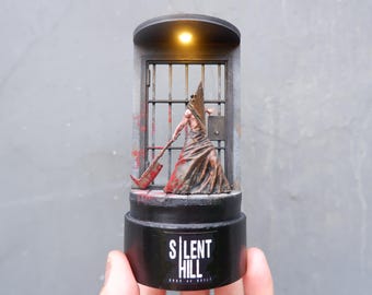 May include: A small, lit, black and silver diorama of a figure from the video game Silent Hill. The figure is holding a large, rusty, bladed weapon. The figure is standing in front of a barred door. The base of the diorama is black and has the words "Silent Hill" printed on it.