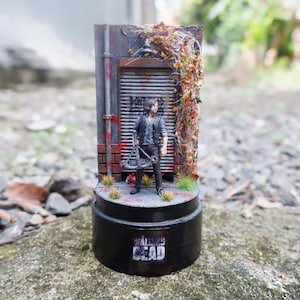 Puede incluir: Figura en miniatura de un personaje de "The Walking Dead" frente a una fachada de edificio desgastada. La base es negra con el logo de la serie. La escena incluye una ballesta y follaje.