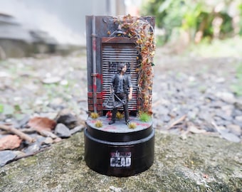 May include: A miniature figurine of a character from "The Walking Dead" stands in front of a weathered building facade. The base is black with the show's logo. The scene includes a crossbow and foliage.