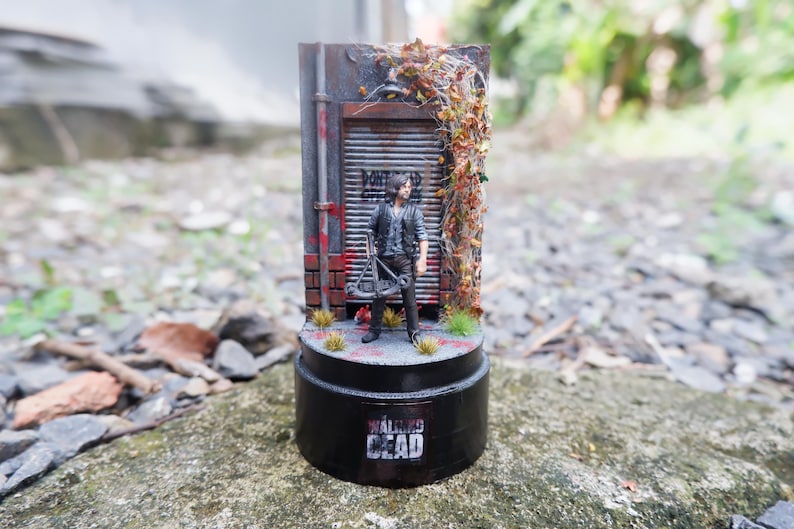 Puede incluir: Figura en miniatura de un personaje de "The Walking Dead" frente a una fachada de edificio desgastada. La base es negra con el logo de la serie. La escena incluye una ballesta y follaje.
