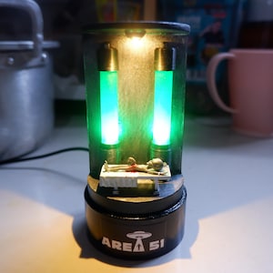 May include: A novelty lamp with an alien autopsy scene. The lamp has two vertical green glowing tubes and a small alien figure on a table. The black base has the words "AREA 51" and a UFO graphic.