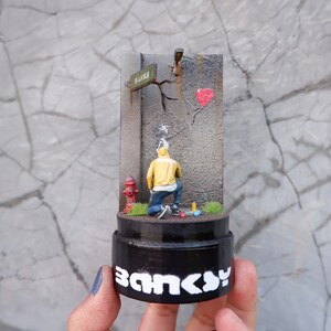 May include: A miniature replica of a famous street art piece by Banksy. The sculpture depicts a hooded figure spray painting a red heart balloon on a grey wall. The sculpture is on a black base with the word "Banksy" written in white letters.