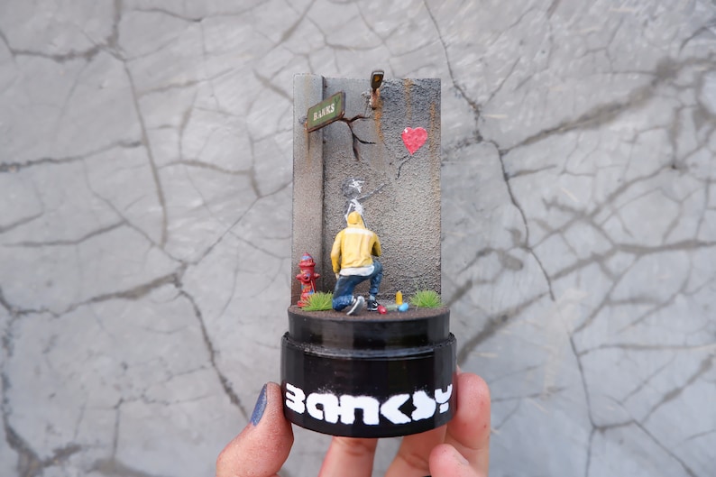 May include: A miniature replica of a famous street art piece by Banksy. The sculpture depicts a hooded figure spray painting a red heart balloon on a grey wall. The sculpture is on a black base with the word "Banksy" written in white letters.