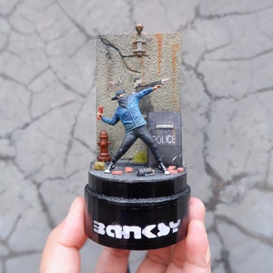May include: A small, detailed figurine of a masked figure throwing a Molotov cocktail. The figure is standing in front of a brick wall with a fire hydrant and a police shield. The base of the figurine is black and says "Banksy" in white letters.