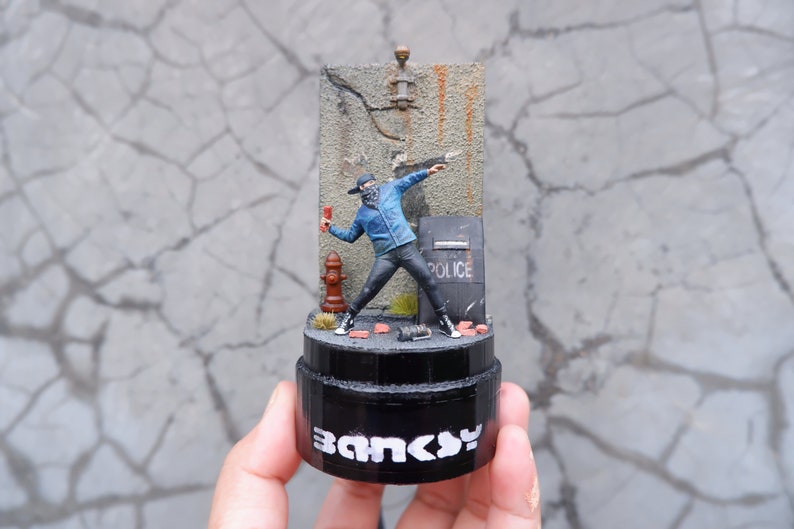May include: A small, detailed figurine of a masked figure throwing a Molotov cocktail. The figure is standing in front of a brick wall with a fire hydrant and a police shield. The base of the figurine is black and says "Banksy" in white letters.