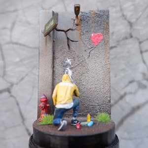 May include: A miniature diorama depicting a street scene with a person in a yellow jacket painting a heart-shaped balloon on a cracked wall. The diorama includes a fire hydrant, a street sign that reads "Banks", and a small patch of green grass.