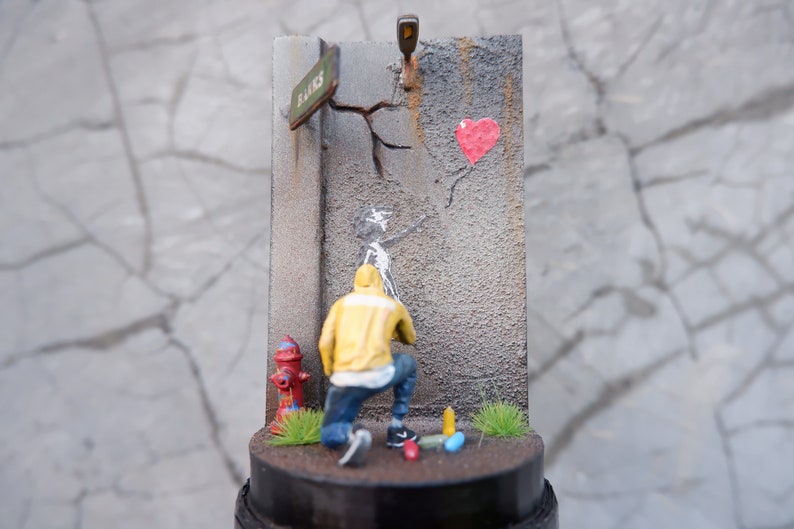 May include: A miniature diorama depicting a street scene with a person in a yellow jacket painting a heart-shaped balloon on a cracked wall. The diorama includes a fire hydrant, a street sign that reads "Banks", and a small patch of green grass.