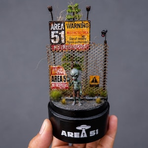 Op de afbeelding: Een miniatuur diorama met een grijze alien-figuur voor een gaashekwerk met verweerde Area 51-waarschuwingsborden. Het geheel is gemonteerd op een zwarte cilindrische voet met de tekst Area 51 en een afbeelding van een vliegende schotel.