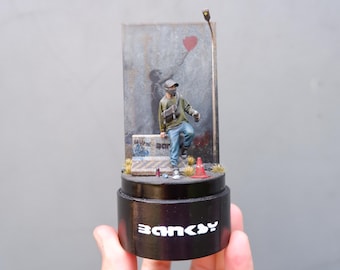 May include: A miniature diorama featuring a figure in a green jacket and black mask, sitting on a concrete barrier with graffiti. The figure is holding a spray can. The diorama is set against a backdrop of a brick wall with a red balloon and a streetlight. The base of the diorama is black and has the word "Banksy" written on it.