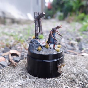 Op de afbeelding: Miniatuurbeeldje van een figuur met bijlen, staande bij een galg. De figuur staat op een rotsachtige basis met gras en een zwarte cilindrische basis. De galg is bruin en de figuur draagt blauwe en bruine kleding.