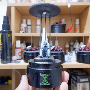 May include: A gray figure sitting in a cage with a black base and a black disc on top. The base has the text "X-Files" printed on it in green.