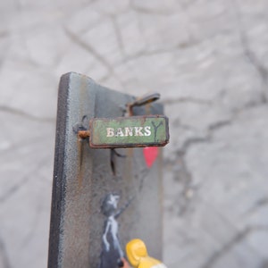 May include: A green metal sign with the word "Banks" written in white letters. The sign is attached to a metal surface with a rusty nail.