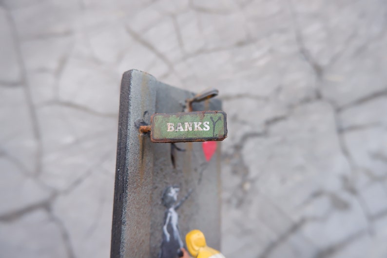May include: A green metal sign with the word "Banks" written in white letters. The sign is attached to a metal surface with a rusty nail.