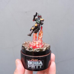 May include: A black and gold Star Wars The Book of Boba Fett collectible figure of Boba Fett in a jetpack with flames.