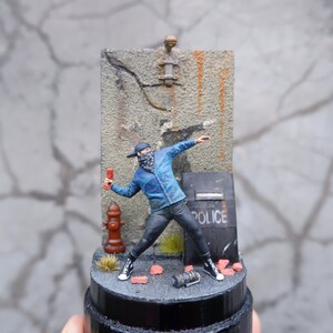 May include: A miniature figure of a person wearing a blue jacket, black pants, and a black face mask, throwing a red object. The figure is standing in front of a gray wall with a police shield and a fire hydrant. The figure is on a black base with red bricks and a black spray can.