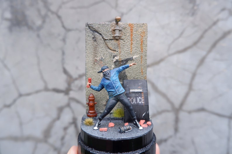 May include: A miniature figure of a person wearing a blue jacket, black pants, and a black face mask, throwing a red object. The figure is standing in front of a gray wall with a police shield and a fire hydrant. The figure is on a black base with red bricks and a black spray can.