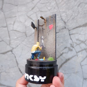 May include: A miniature diorama showing a person in a yellow hoodie spray painting a stencil on a cracked wall with a red heart painted on it. The diorama is on a black base with the word "INCY" written on it.