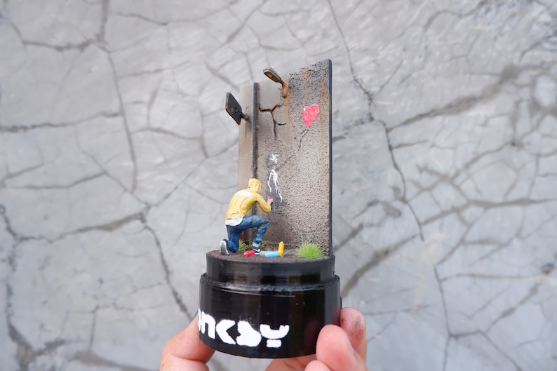 May include: A miniature diorama showing a person in a yellow hoodie spray painting a stencil on a cracked wall with a red heart painted on it. The diorama is on a black base with the word "INCY" written on it.