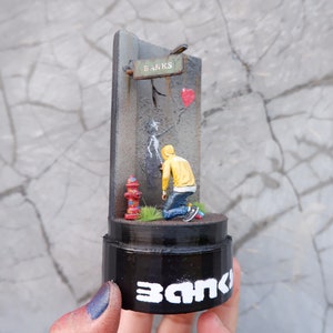 May include: A miniature 3D sculpture of a person in a yellow hoodie spray painting a wall with a red heart and the word "Banks" on a sign above. The sculpture is on a black base with the word "Banks" written on it.