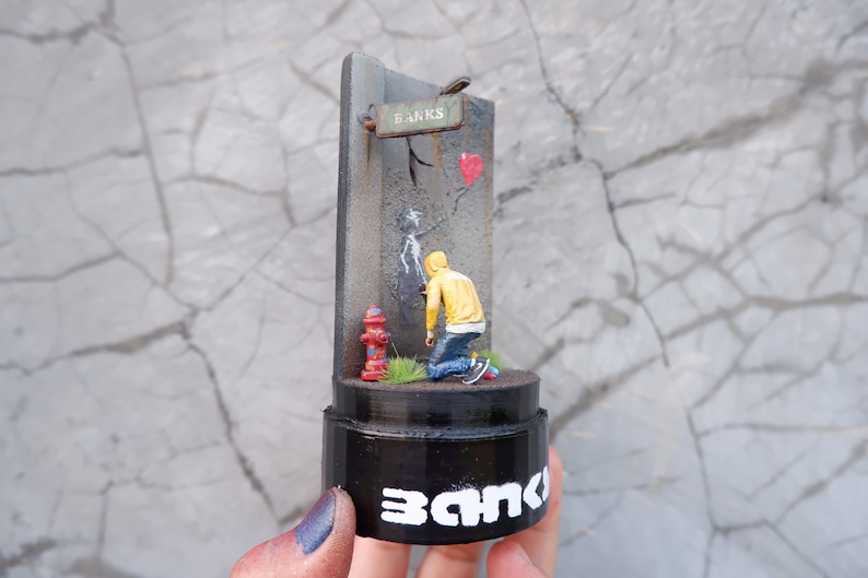 May include: A miniature 3D sculpture of a person in a yellow hoodie spray painting a wall with a red heart and the word "Banks" on a sign above. The sculpture is on a black base with the word "Banks" written on it.