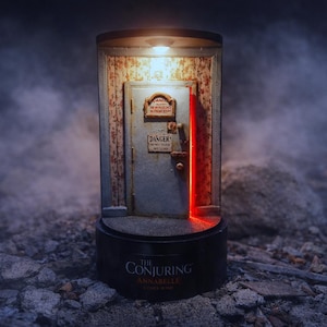 May include: A miniature diorama of a gray door set within a frame, featuring two signs that read DANGER and DO NOT OPEN. The door is illuminated by a top light, casting a red glow from the side, and sits on a black base labeled THE CONJURING ANNABELLE.