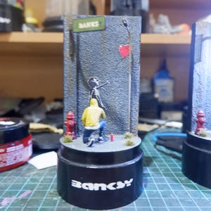 May include: A miniature replica of a Banksy street art scene. The scene features a figure in a yellow jacket spray painting a stencil of a girl holding a red balloon on a grey brick wall. The scene is set on a black base with the word "Banksy" printed in white.