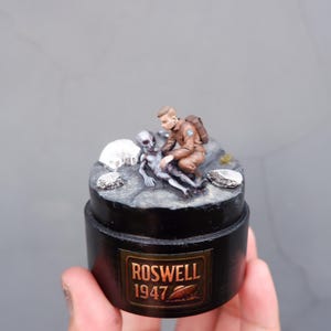 Pode incluir: Diorama em miniatura retratando uma cena de Roswell, 1947. Apresenta uma figura em uniforme marrom cuidando de um alienígena cinza em uma superfície rochosa. A base preta tem um rótulo dourado que diz "Roswell 1947".