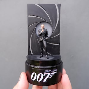 Pode incluir: Uma figura de James Bond em preto e prata em pé em frente a um design de alvo em preto e branco. A base da figura é preta e tem o texto "JAMES BOND 007" impresso nela.