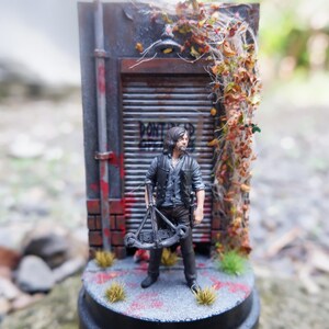 Puede incluir: Figura en miniatura de un hombre con una ballesta, frente a una fachada de edificio desgastada. La escena incluye una pared de ladrillo, una puerta met&aacute;lica con graffiti y enredaderas. La base es una plataforma circular.
