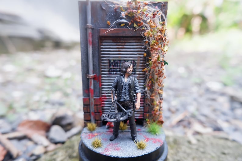 Puede incluir: Figura en miniatura de un hombre con una ballesta, frente a una fachada de edificio desgastada. La escena incluye una pared de ladrillo, una puerta met&aacute;lica con graffiti y enredaderas. La base es una plataforma circular.