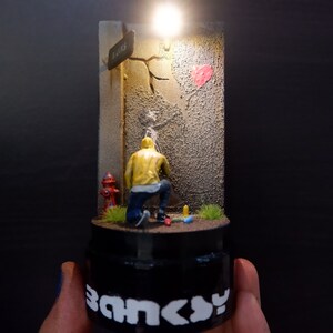 May include: A miniature diorama depicting a street scene with a figure painting a heart on a wall. The figure is wearing a yellow jacket and blue jeans. The diorama is lit from above and has a black base with the word "Banksy" printed on it.