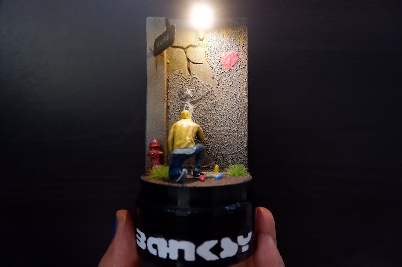 May include: A miniature diorama depicting a street scene with a figure painting a heart on a wall. The figure is wearing a yellow jacket and blue jeans. The diorama is lit from above and has a black base with the word "Banksy" printed on it.
