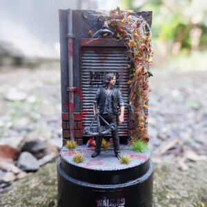 Puede incluir: Figura en miniatura de un personaje de "The Walking Dead" frente a un tel&oacute;n de fondo desgastado. La escena incluye una puerta de metal, ladrillos y follaje. La base presenta el logotipo de la serie. La figura sostiene una ballesta.
