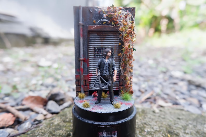 Puede incluir: Figura en miniatura de un personaje de "The Walking Dead" frente a un tel&oacute;n de fondo desgastado. La escena incluye una puerta de metal, ladrillos y follaje. La base presenta el logotipo de la serie. La figura sostiene una ballesta.