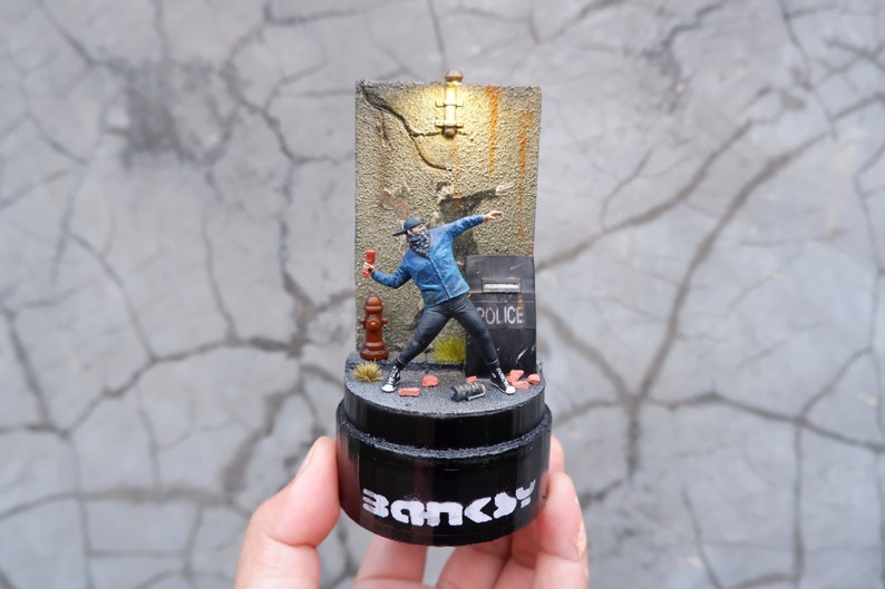 May include: A small, detailed figurine of a masked figure throwing a fire extinguisher at a wall with the word "Police" on it. The figurine is on a black base with the word "Banksy" printed on it.