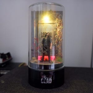Puede incluir: L&aacute;mpara decorativa cil&iacute;ndrica con una figura de la serie "The Walking Dead". La figura est&aacute; frente a una puerta met&aacute;lica, iluminada por una luz c&aacute;lida. La base de la l&aacute;mpara es negra con el t&iacute;tulo de la serie en blanco.