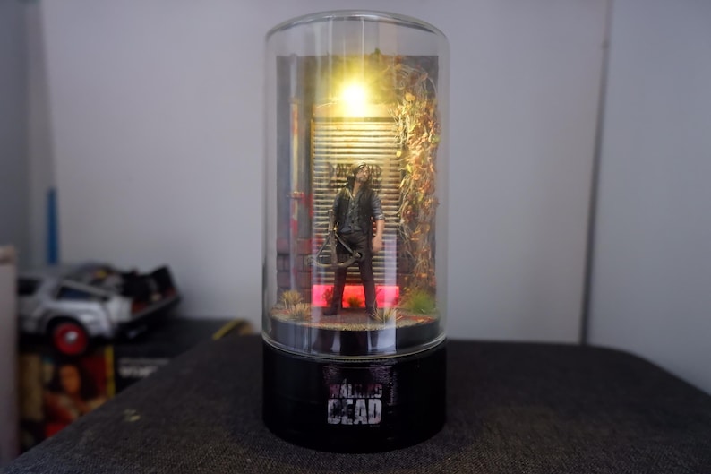 Puede incluir: L&aacute;mpara decorativa cil&iacute;ndrica con una figura de la serie "The Walking Dead". La figura est&aacute; frente a una puerta met&aacute;lica, iluminada por una luz c&aacute;lida. La base de la l&aacute;mpara es negra con el t&iacute;tulo de la serie en blanco.