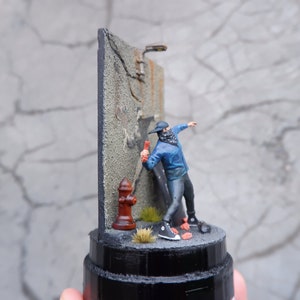 May include: A miniature figurine of a person wearing a blue jacket and black pants spray painting a wall. The wall is made of gray concrete and has a red fire hydrant at the base. The figurine is standing on a black base.